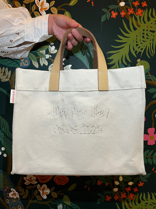 A white canvas tote with leather handles and embroidered text 'Wifey for Lifey' along with a date '05.05.2024' on the front.