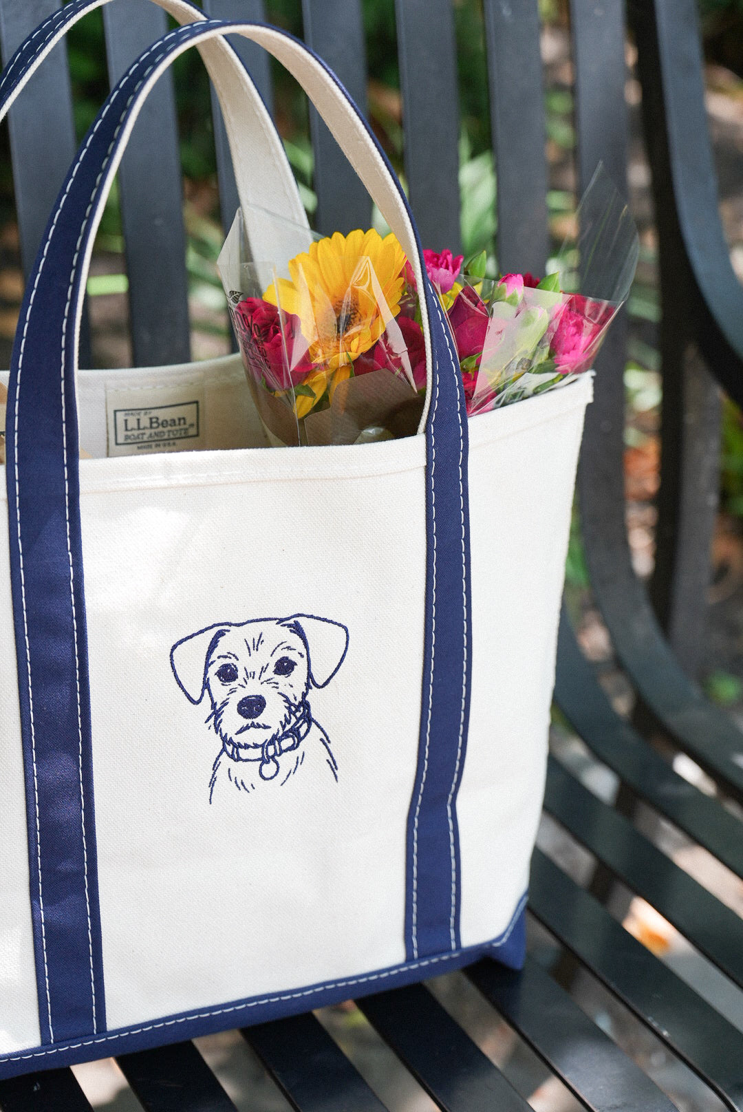 White tote bag with navy handles and an embroidered dog portrait, carrying a bouquet of flowers