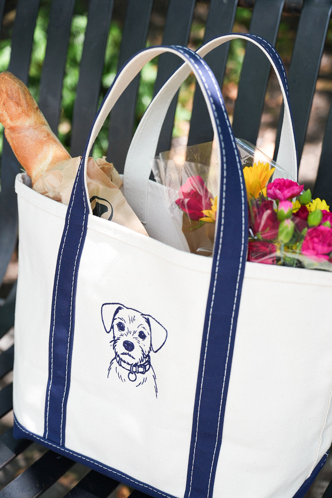 White tote bag with navy straps and an embroidered dog portrait, carrying flowers and bread
