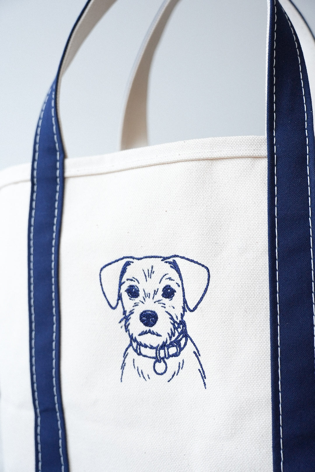 Close-up of a white tote bag with navy blue straps and an embroidered dog portrait