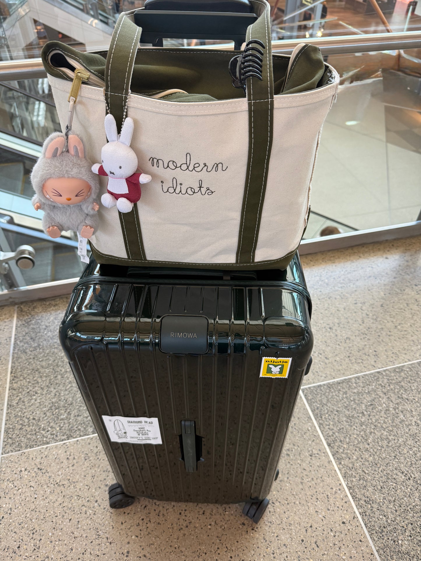A white and olive green tote bag with embroidered text labelled 'modern idiots', placed on top of a black suitcase.
