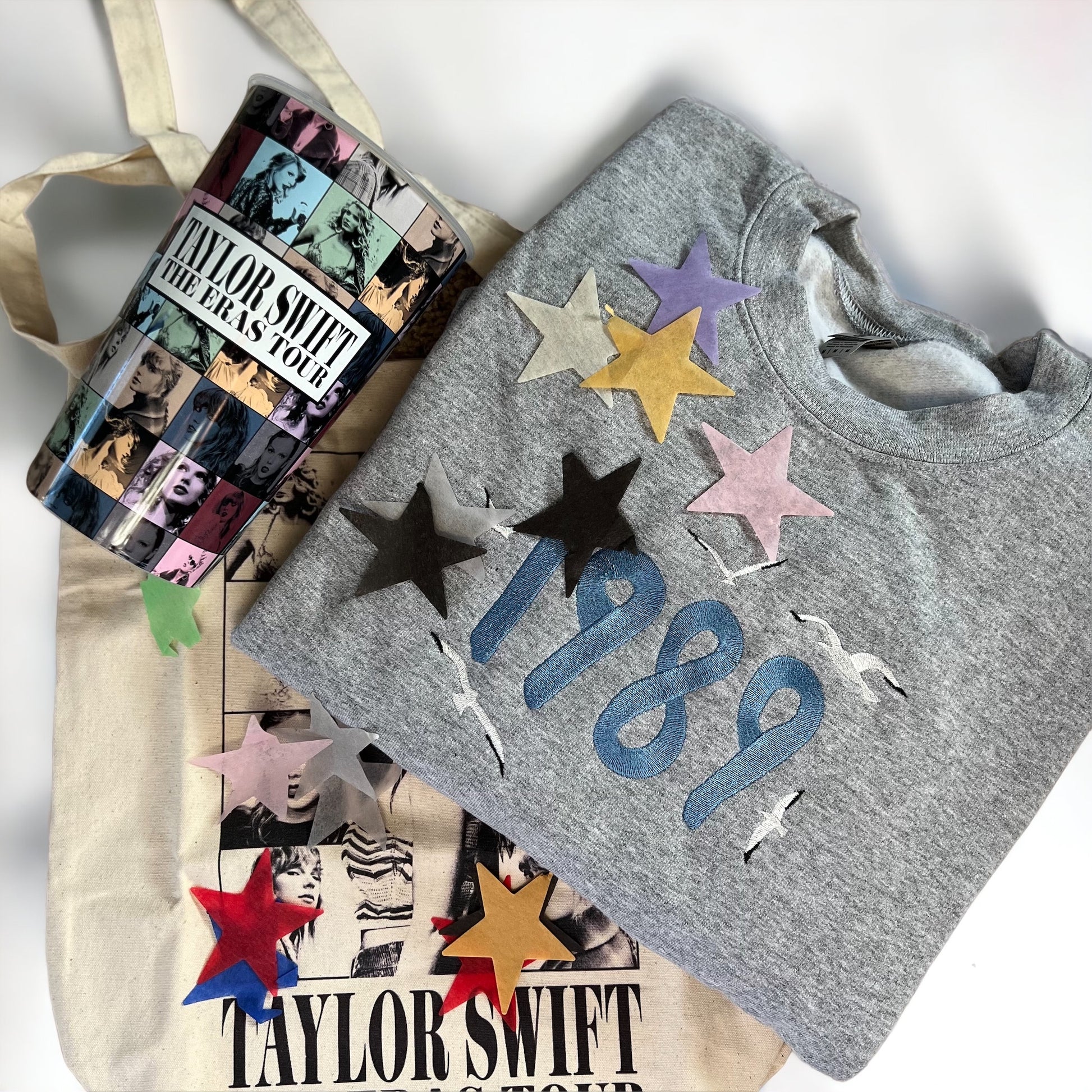 A gray sweatshirt with embroidered "1989" text and seagulls on the front, placed next to Taylor Swift Eras Tour merchandise