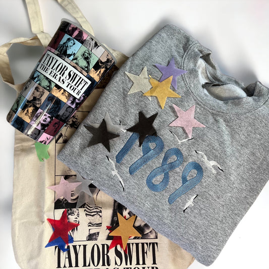 A gray sweatshirt with embroidered "1989" text and seagulls on the front, placed next to Taylor Swift Eras Tour merchandise
