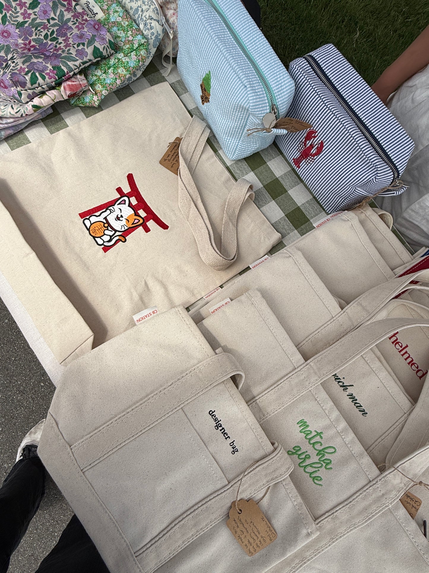 Beige tote bag with an embroidered Maneki Neko Torii design, displayed beside other embroidered designs