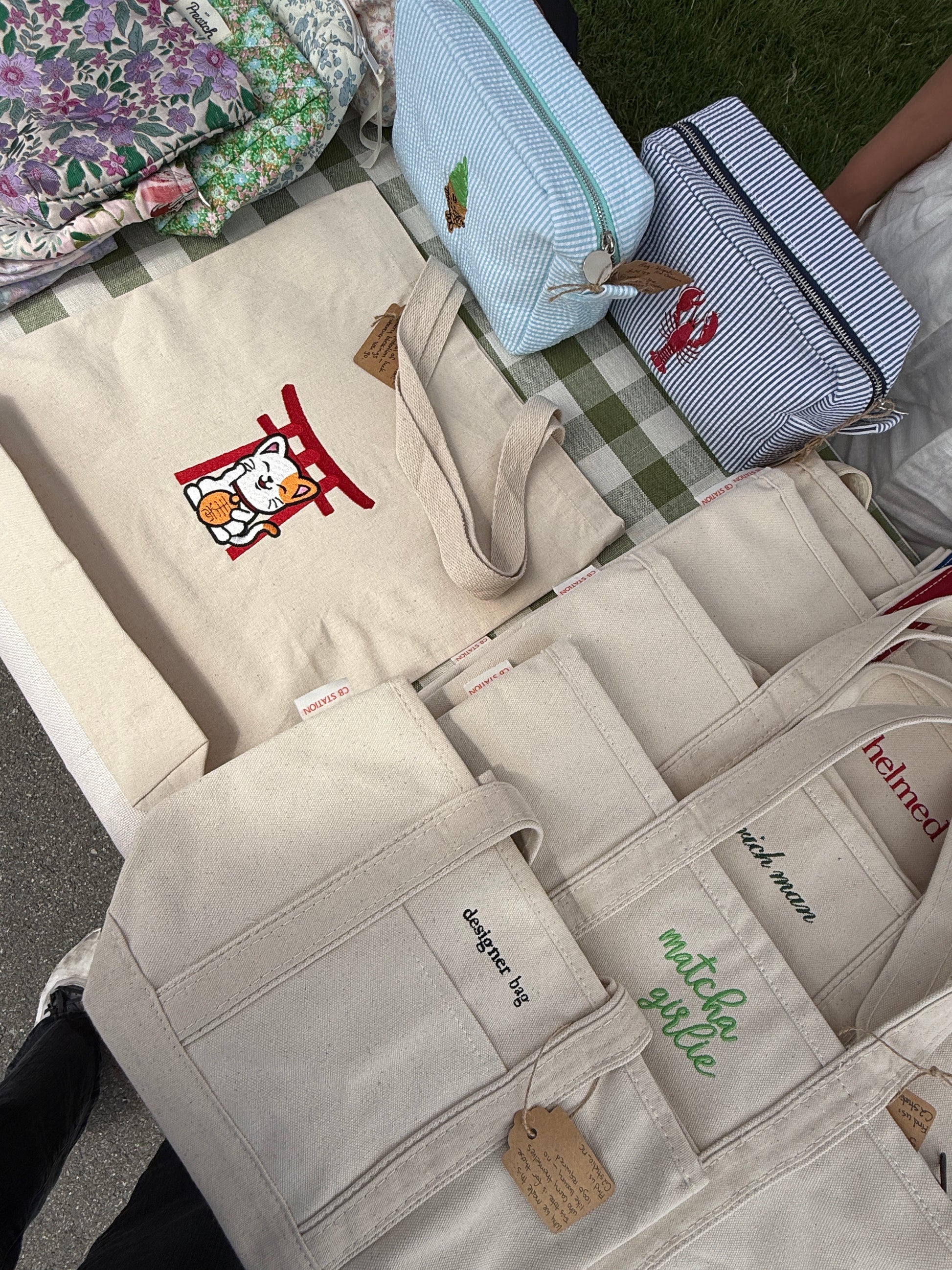 Beige tote bag with an embroidered Maneki Neko Torii design, displayed beside other embroidered designs