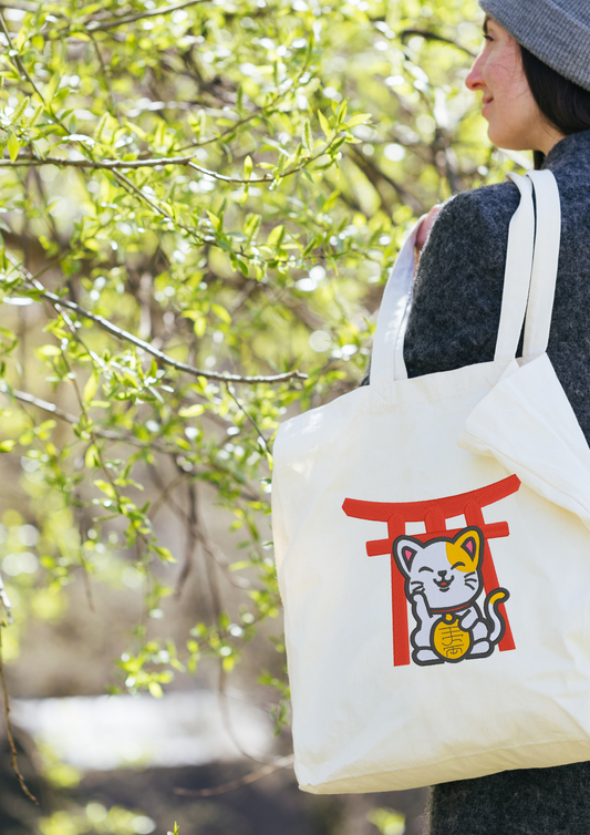 Person holding a beige tote bag with an embroidered Maneki Neko Torii design