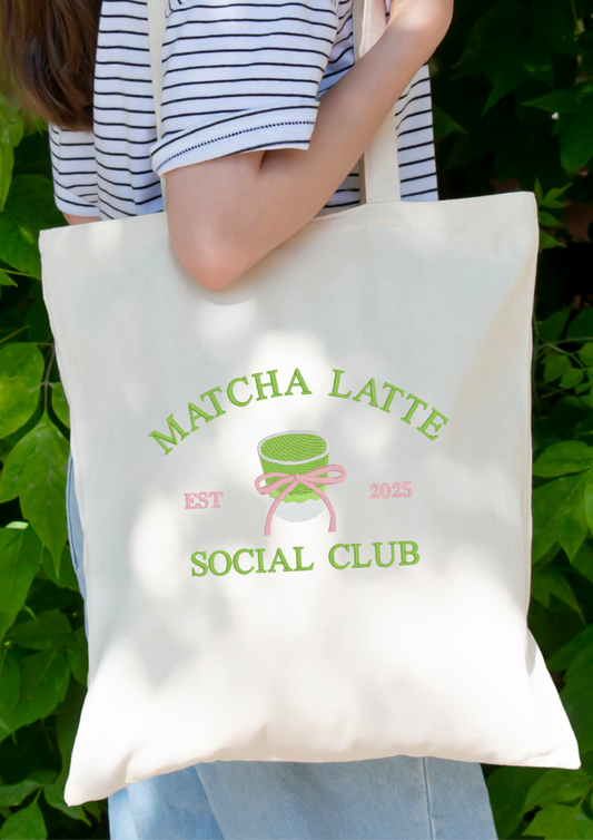 Person holding a beige tote bag with an embroidered 'Matcha Latte Social Club EST 2025' design