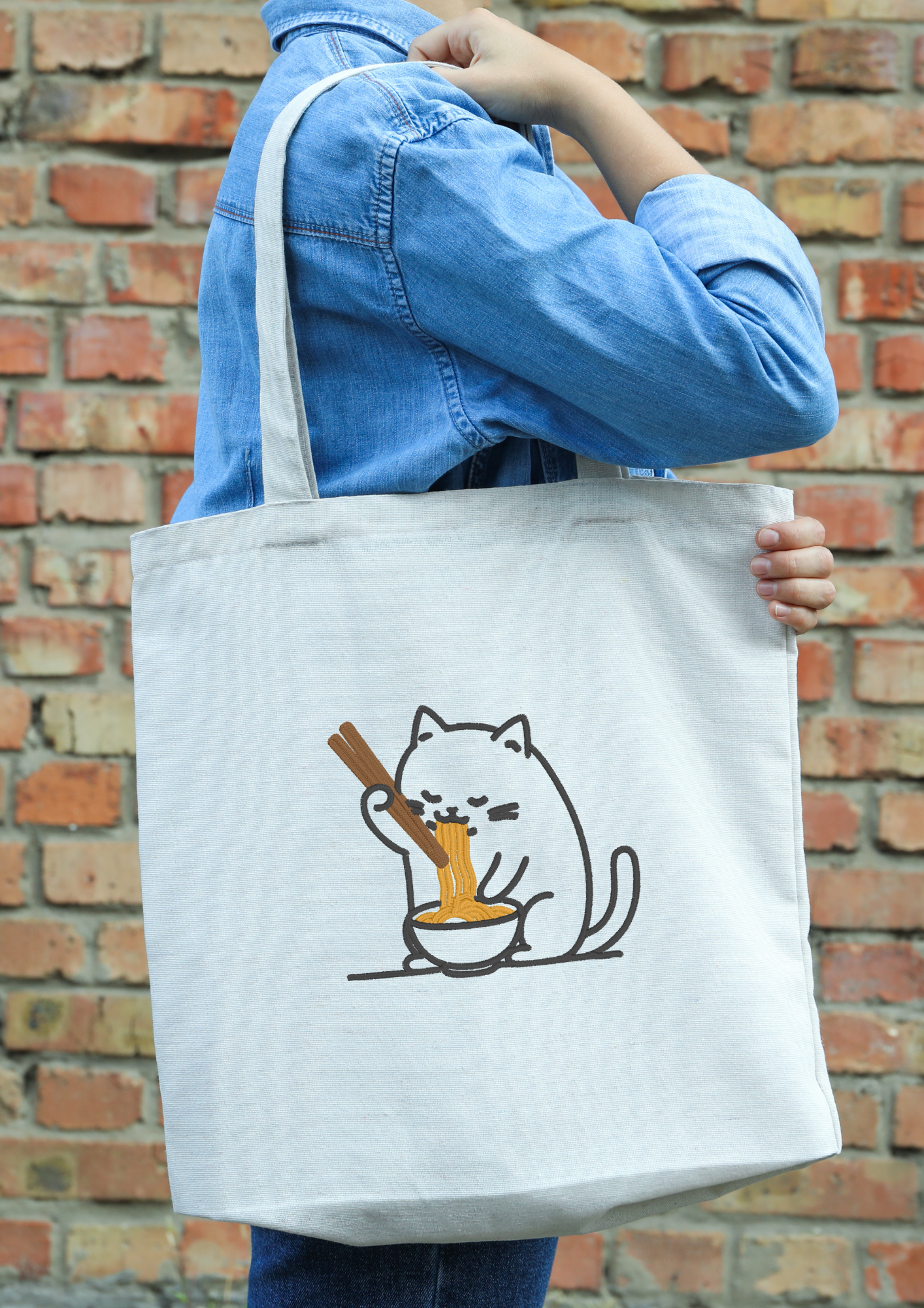 Person holding a beige tote bag with an embroidered Slurping Ramen Cat design
