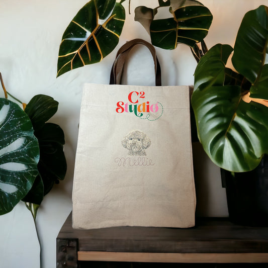 A beige canvas tote bag with leather handles featuring an embroidered illustration of a dog portrait.